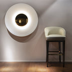 A white Clérachic wall light glows softly near a cream barstool, its brass centre warm against the pale shade.