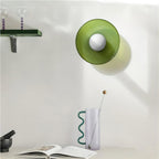 A green cylindrical wall lamp beside a vase and open book, a different fixture from this Clérachic ceiling light.