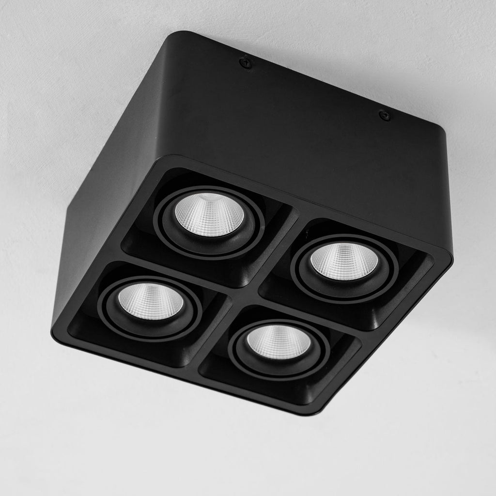 From a closer angle, this black four-light Clérachic spotlight glows warm against a plain white background.