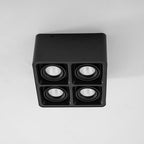 Facing forward, this black four-light Clérachic spotlight glows warm from all four heads on a plain background.