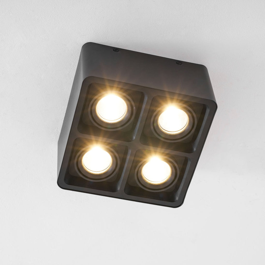 Four warm bulbs glow from this black Clérachic spotlight, angled from below against a plain white background.