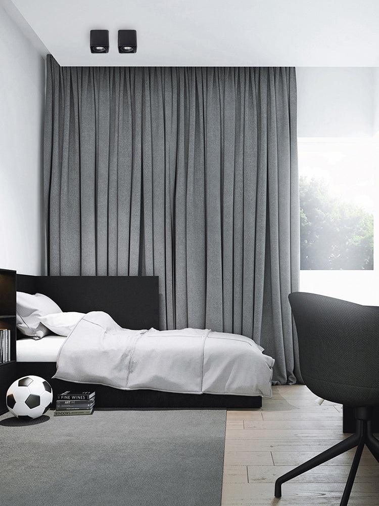 In a bedroom with grey curtains and a dark bed frame, two black Clérachic spotlights sit unlit on the ceiling.