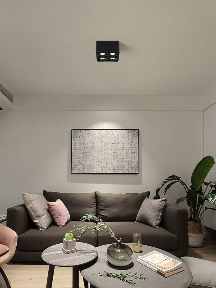 Above a sofa with cushions and a potted plant, a black two-light Clérachic spotlight sits unlit on the ceiling.