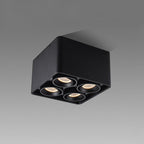 Angled from below-left, this black four-light Clérachic spotlight glows warm against a plain white background.