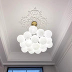 Beneath an ornate ceiling medallion, this Clérachic cluster chandelier hangs unlit, a bright window visible behind it.