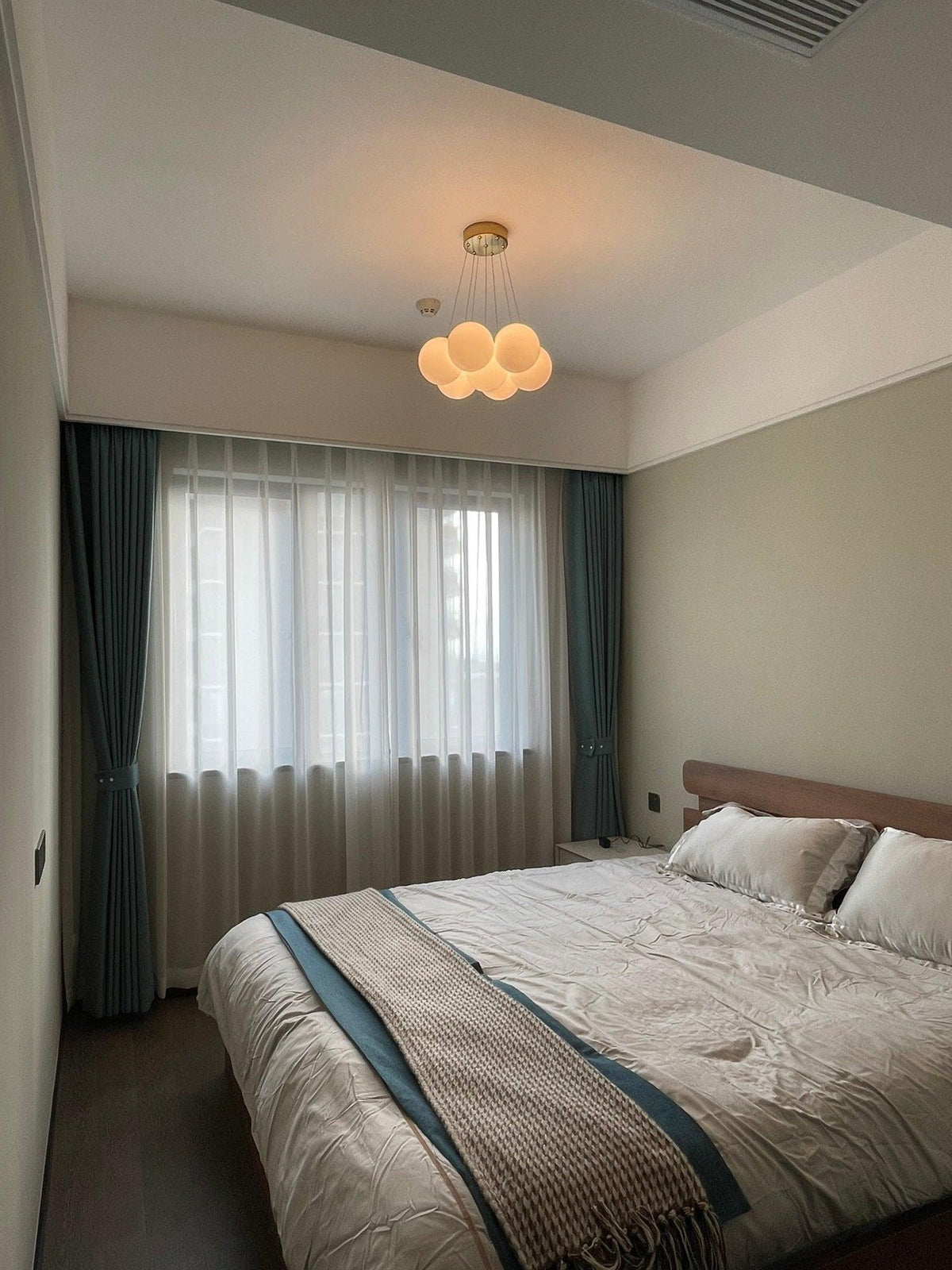 Above a bed dressed in pale linens, this Clérachic cluster chandelier glows softly, sheer curtains at the window.