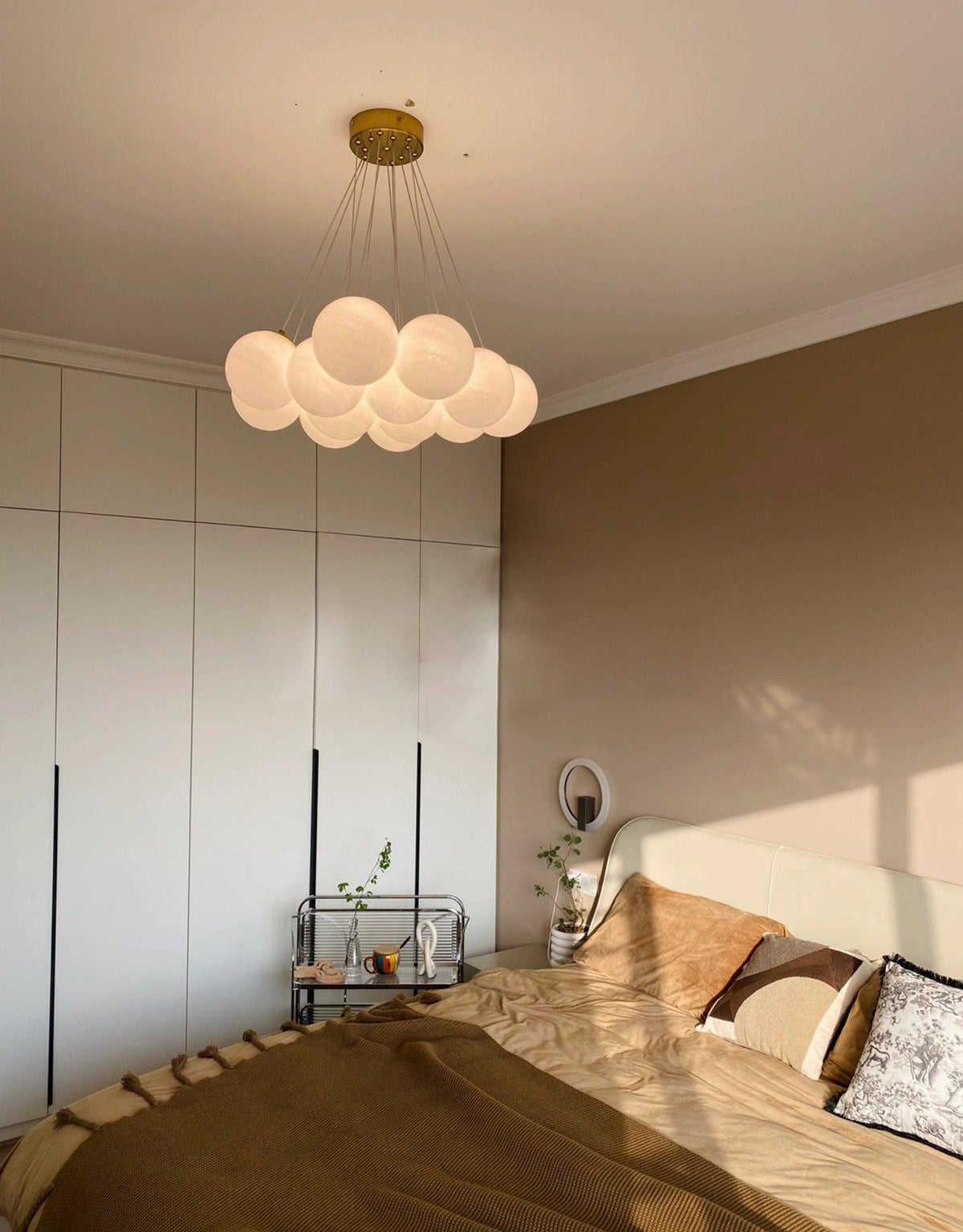 Above a bed in a warm-toned bedroom, this Clérachic cluster chandelier glows beneath sloped wall detailing.