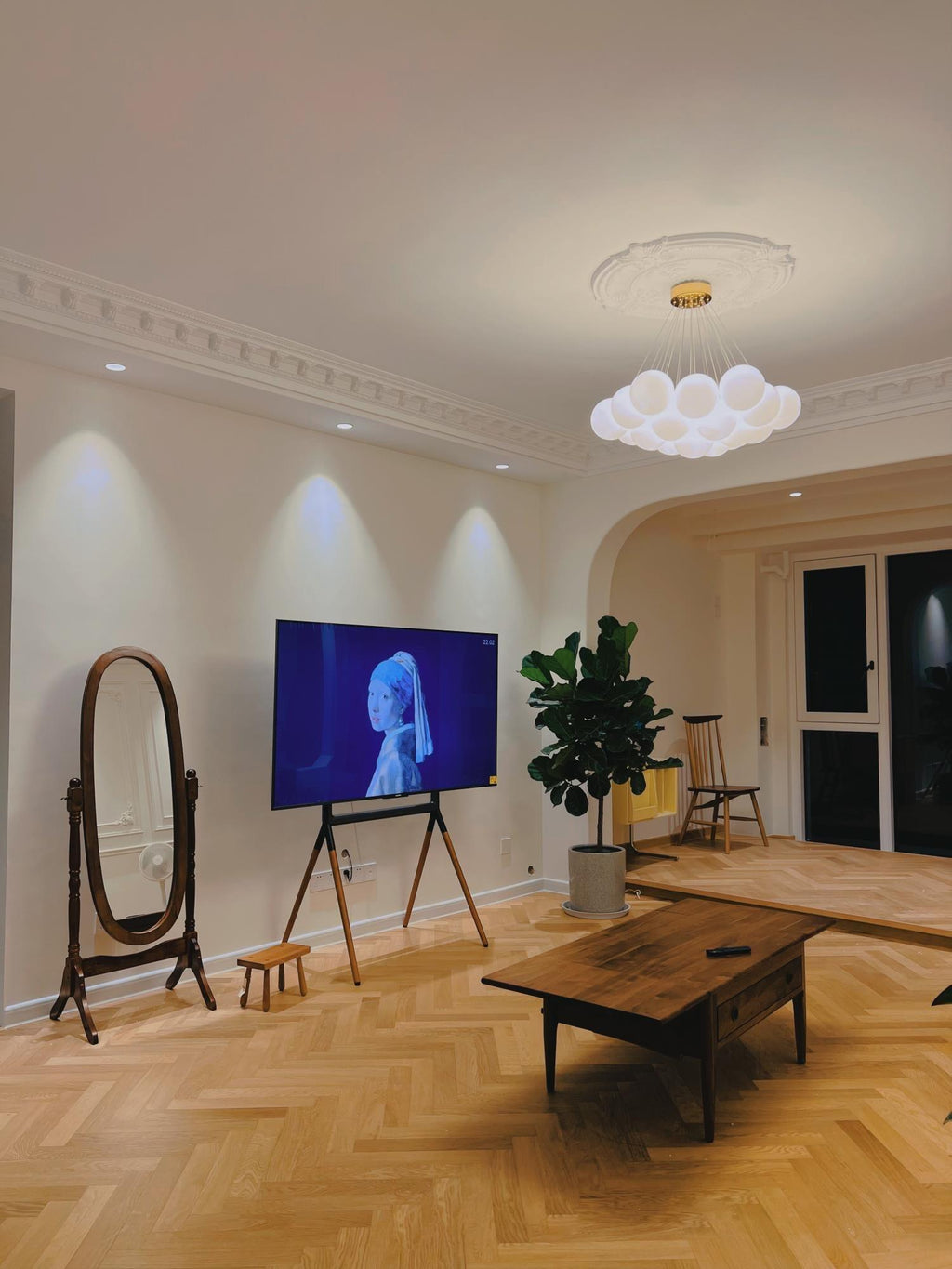 A floor mirror and television sit near this lit Clérachic cluster chandelier above a herringbone floor.