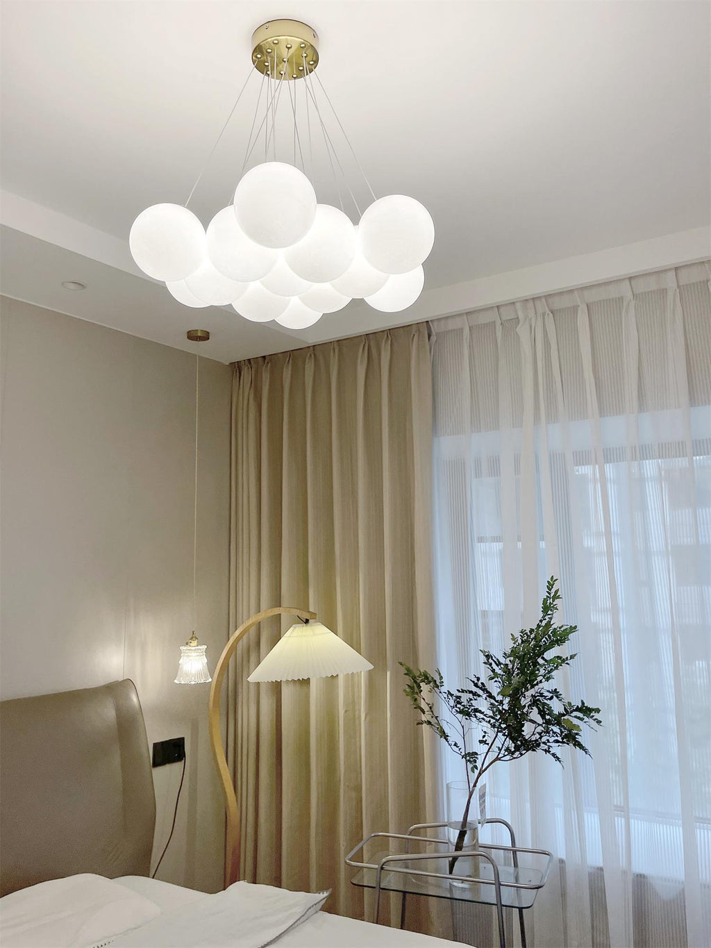 Beside a floor lamp and a small potted plant, this lit Clérachic cluster chandelier hangs in a bedroom.