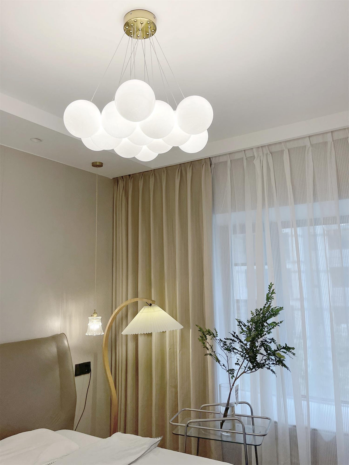 Beside a floor lamp and a small potted plant, this lit Clérachic cluster chandelier hangs in a bedroom.