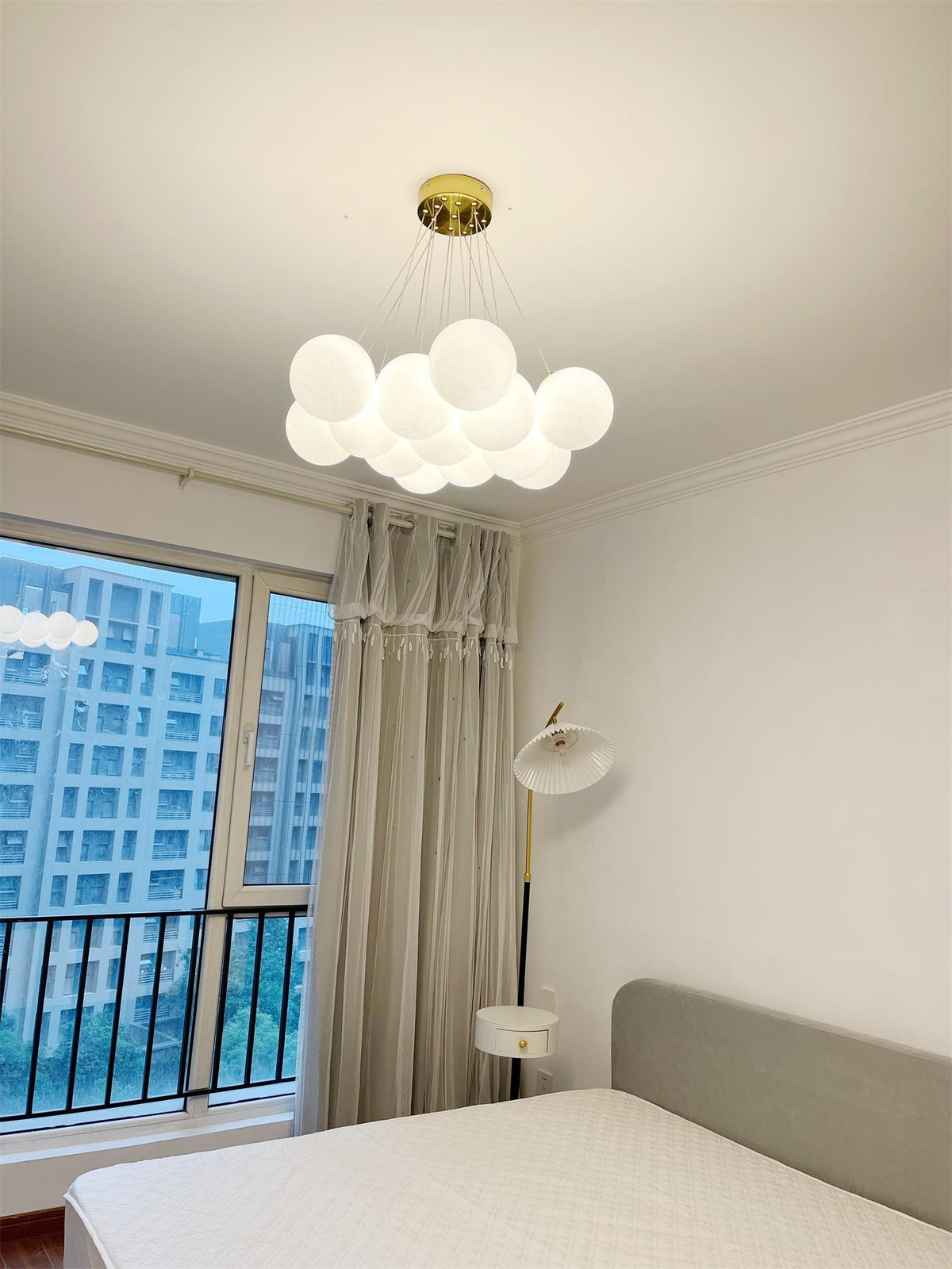 A window overlooking the city sits beside this lit Clérachic cluster chandelier above a grey-dressed bed.