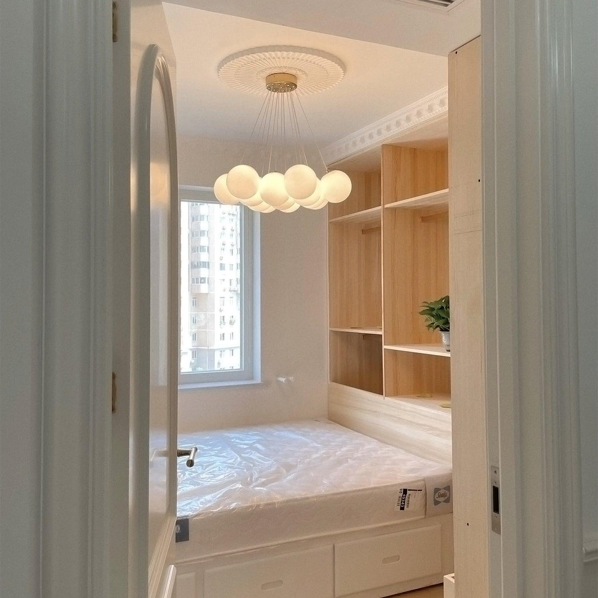 Through a doorway, this Clérachic cluster chandelier hangs unlit above a bed and pale wood cabinetry.