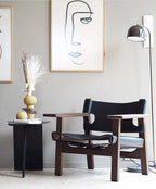 A dark table with framed line art sits near this gold dome-shade Clérachic floor lamp and a black leather chair.
