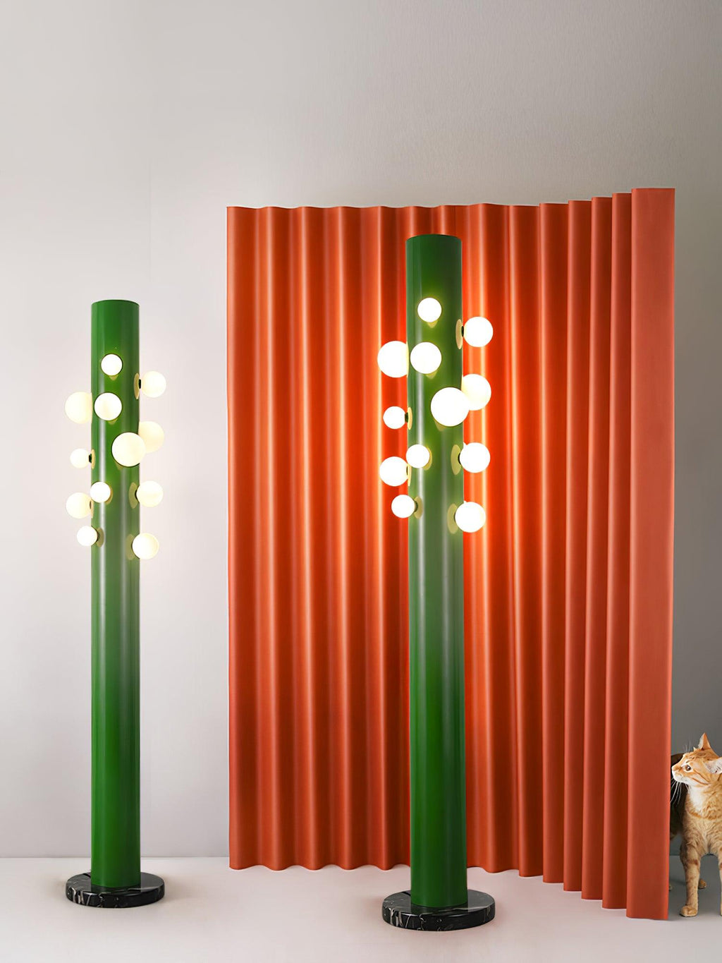 Two green Clérachic floor lamps of different heights stand before a rippled orange backdrop, with a cat resting nearby.