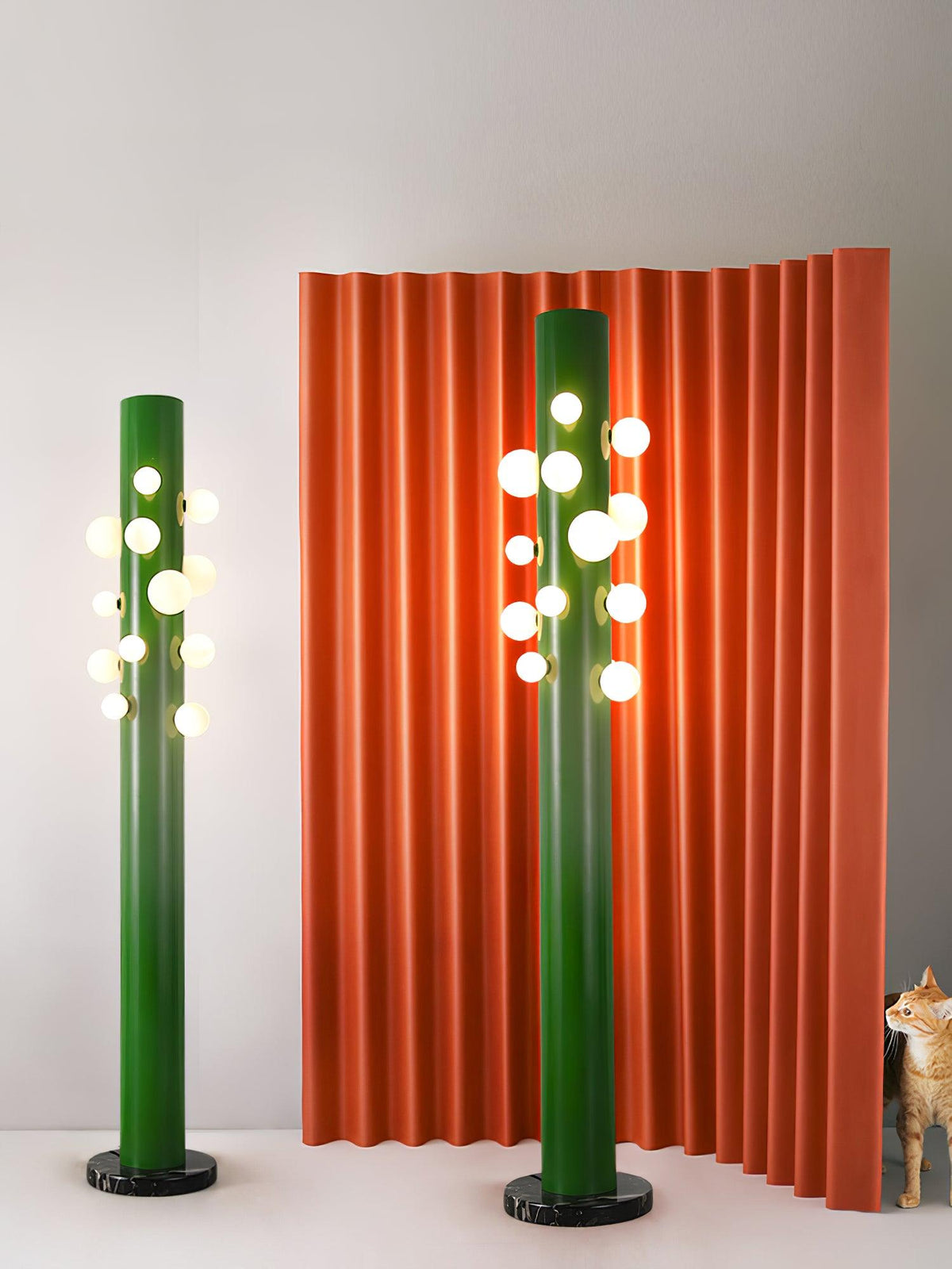 Two green Clérachic floor lamps of different heights stand before a rippled orange backdrop, with a cat resting nearby.