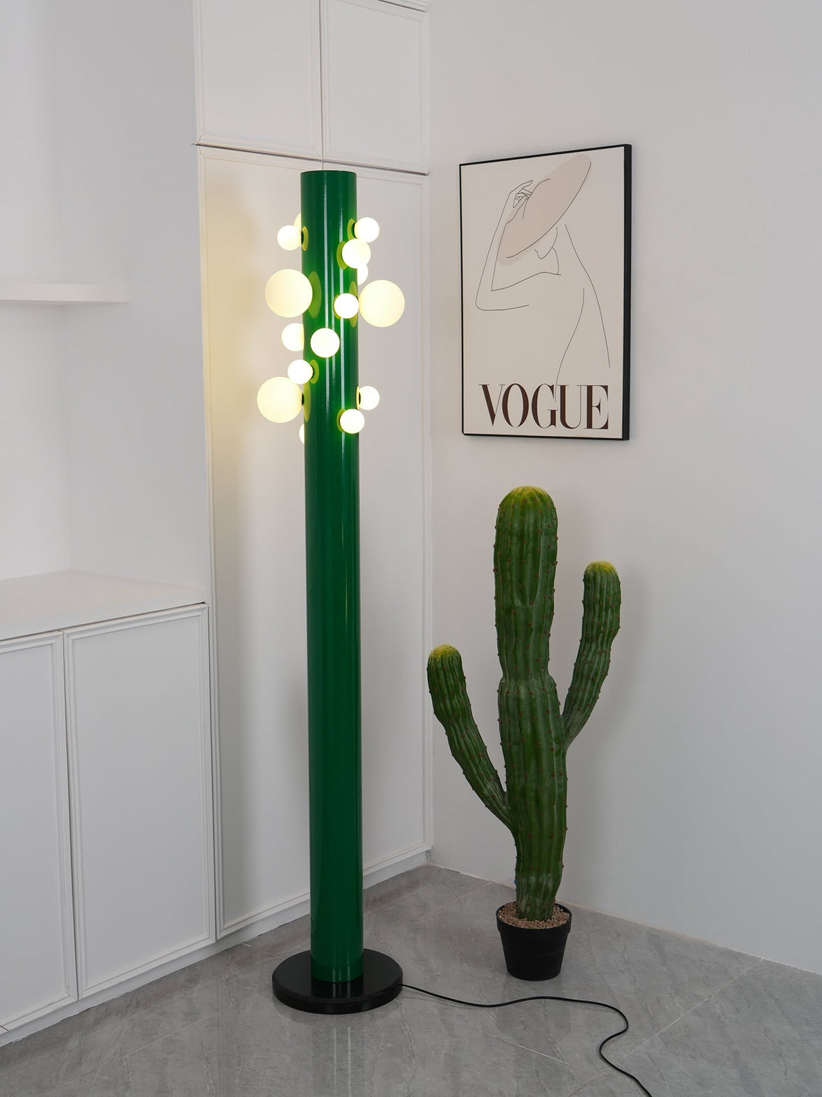 Warm light spills onto the wall as the green Clérachic floor lamp glows brightly beside the potted cactus and framed print.