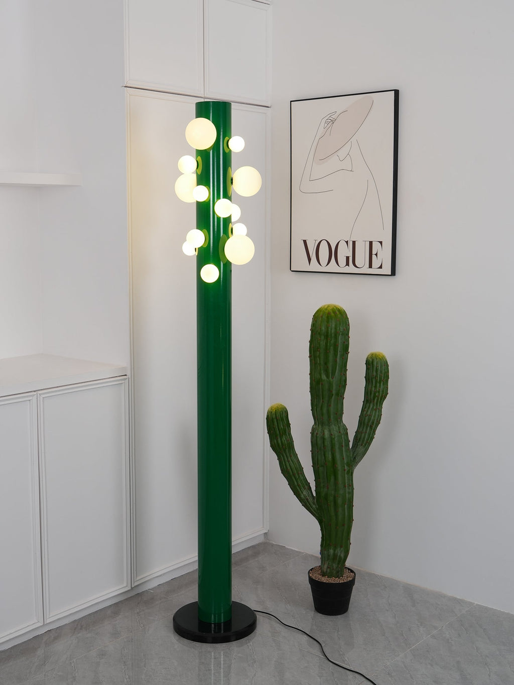 Angled to cast a shadow across the wall, the lit green Clérachic floor lamp stands beside the cactus and fashion print.