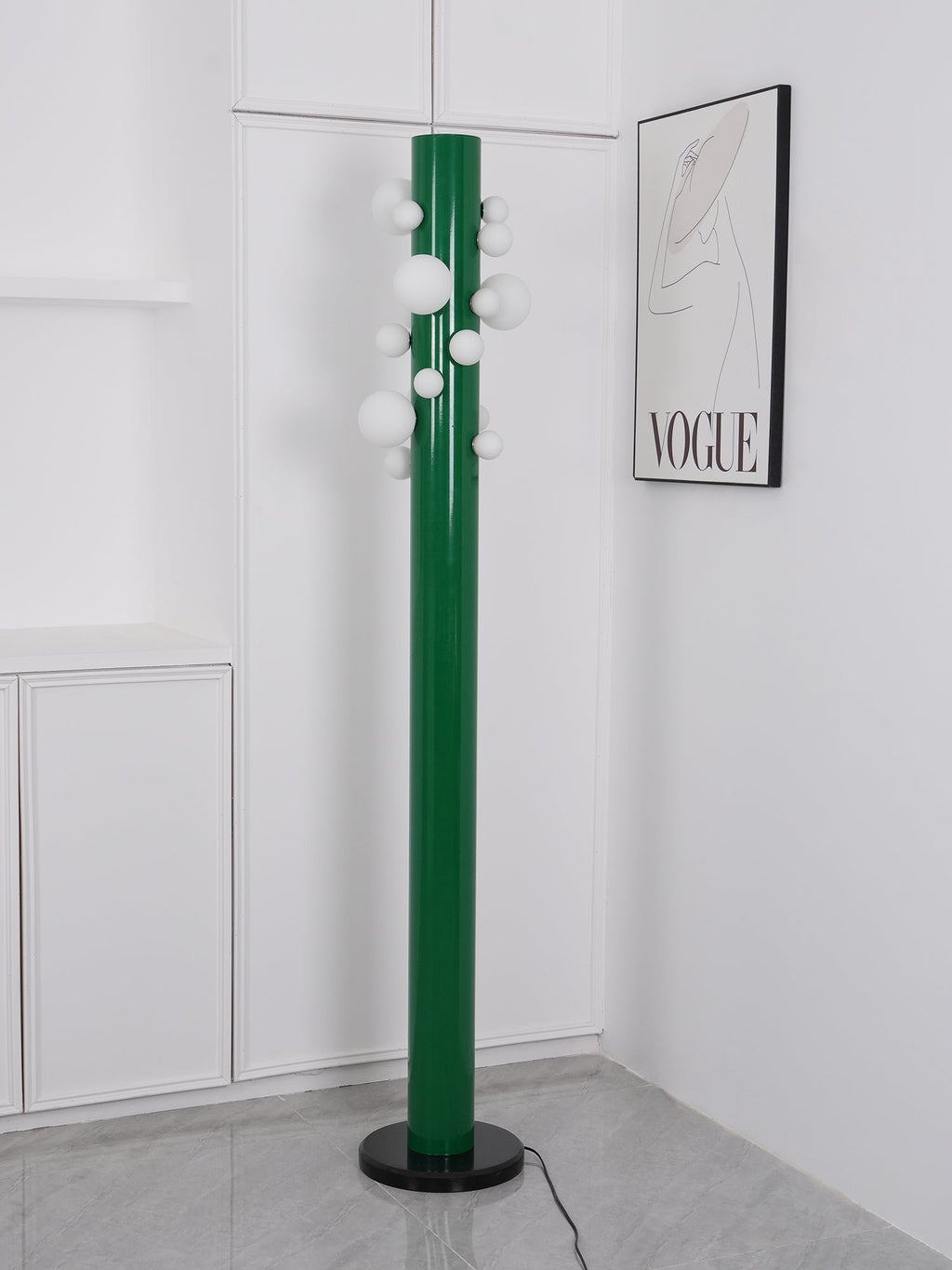 In an all-white corner beneath a monochrome fashion print, the green Clérachic floor lamp's globes glow softly, no plant nearby.