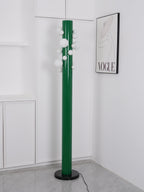 In an all-white corner beneath a monochrome fashion print, the green Clérachic floor lamp's globes glow softly, no plant nearby.
