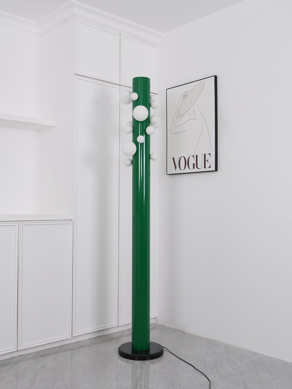 In the same white corner, the green Clérachic floor lamp appears unlit, its frosted globes plain against the fashion print.