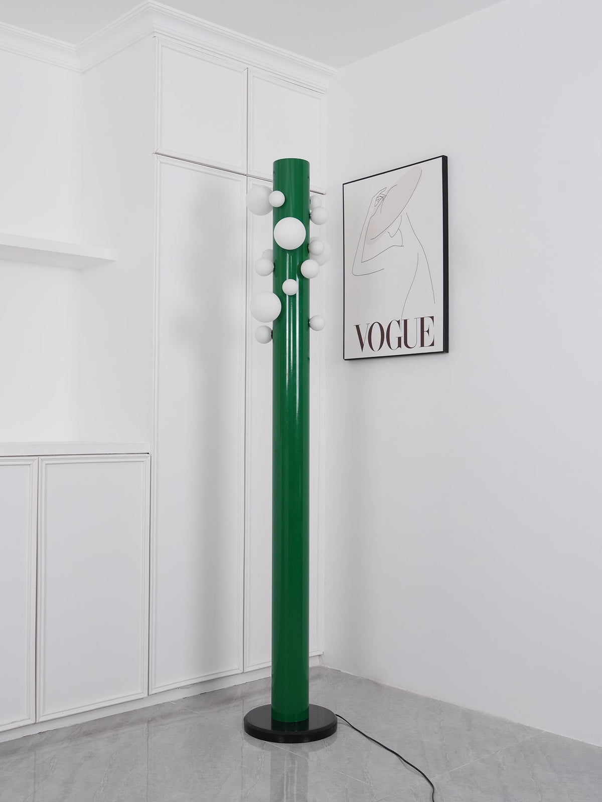 In the same white corner, the green Clérachic floor lamp appears unlit, its frosted globes plain against the fashion print.