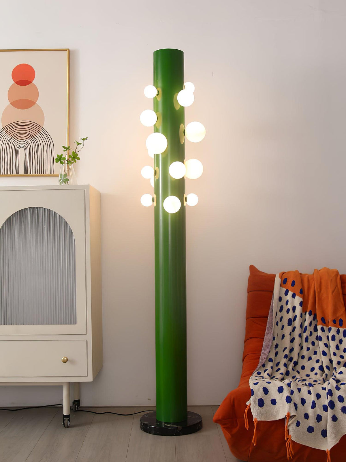 Near a cream cabinet and an orange spotted throw, the green Clérachic floor lamp's glass globes glow above a wood floor.