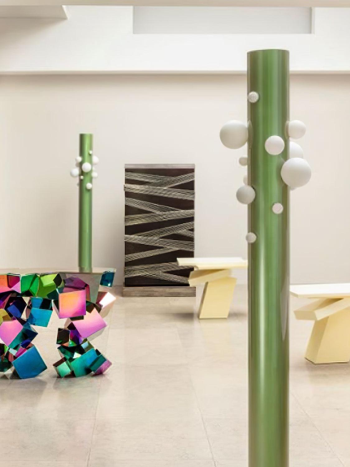 In a gallery-like room with a colourful sculpture and angular furniture, two green Clérachic floor lamps stand at different depths.