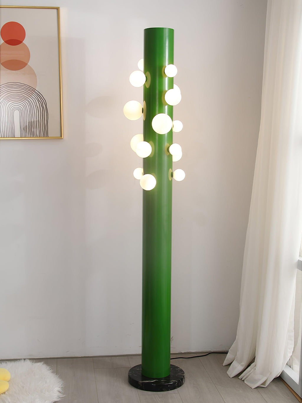 Beside a sheer curtain and a framed abstract print, the green Clérachic floor lamp lights a corner of pale wood flooring.
