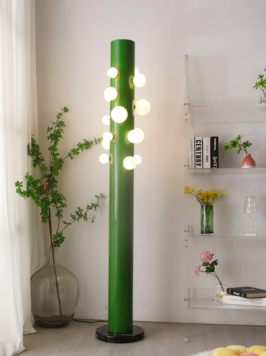 Beside sheer curtains, the green Clérachic floor lamp glows next to a shelf holding books, glassware and a vase of flowers.