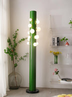 Beside sheer curtains, the green Clérachic floor lamp glows next to a shelf holding books, glassware and a vase of flowers.