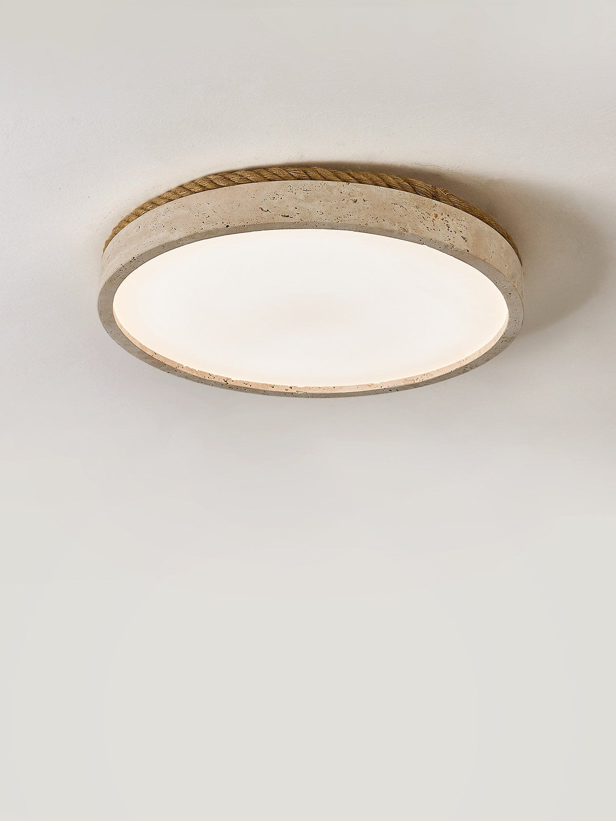This round Clérachic ceiling light, its travertine ring bound in wound jute rope, shown glowing on a plain grey background.
