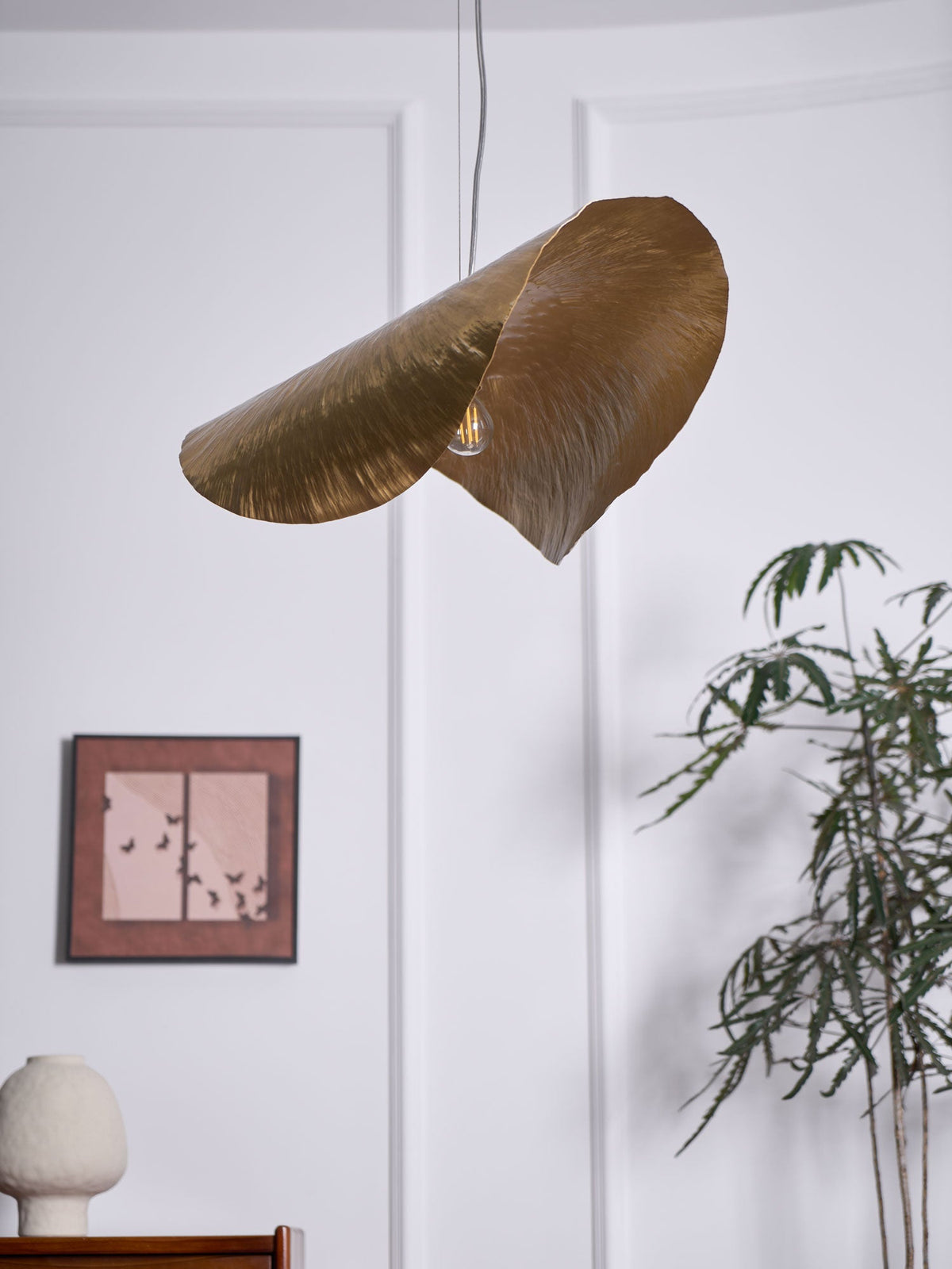 This Clérachic hammered brass shade hangs unlit again beside the framed print and potted plant.