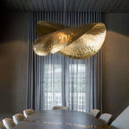 A large hammered brass shade from Clérachic glows above a dining table in a dark, curtained room.