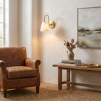 This brushed brass Clérachic wall sconce glows above a wooden bench styled with a vase beside a leather armchair.