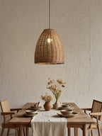 A woven rattan Clérachic pendant glows above a wood table set with dried flowers and rustic chairs.