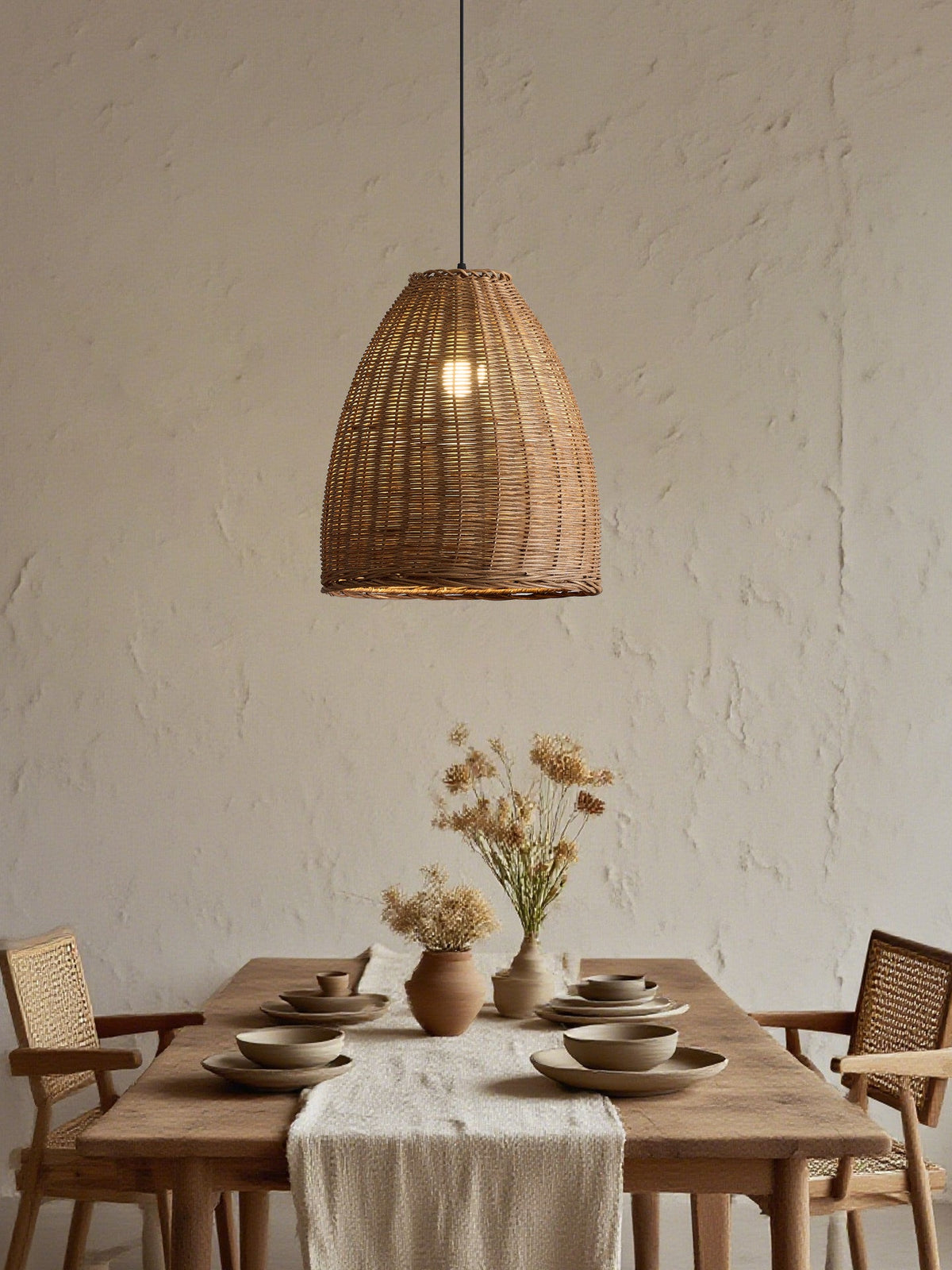 A woven rattan Clérachic pendant glows above a wood table set with dried flowers and rustic chairs.