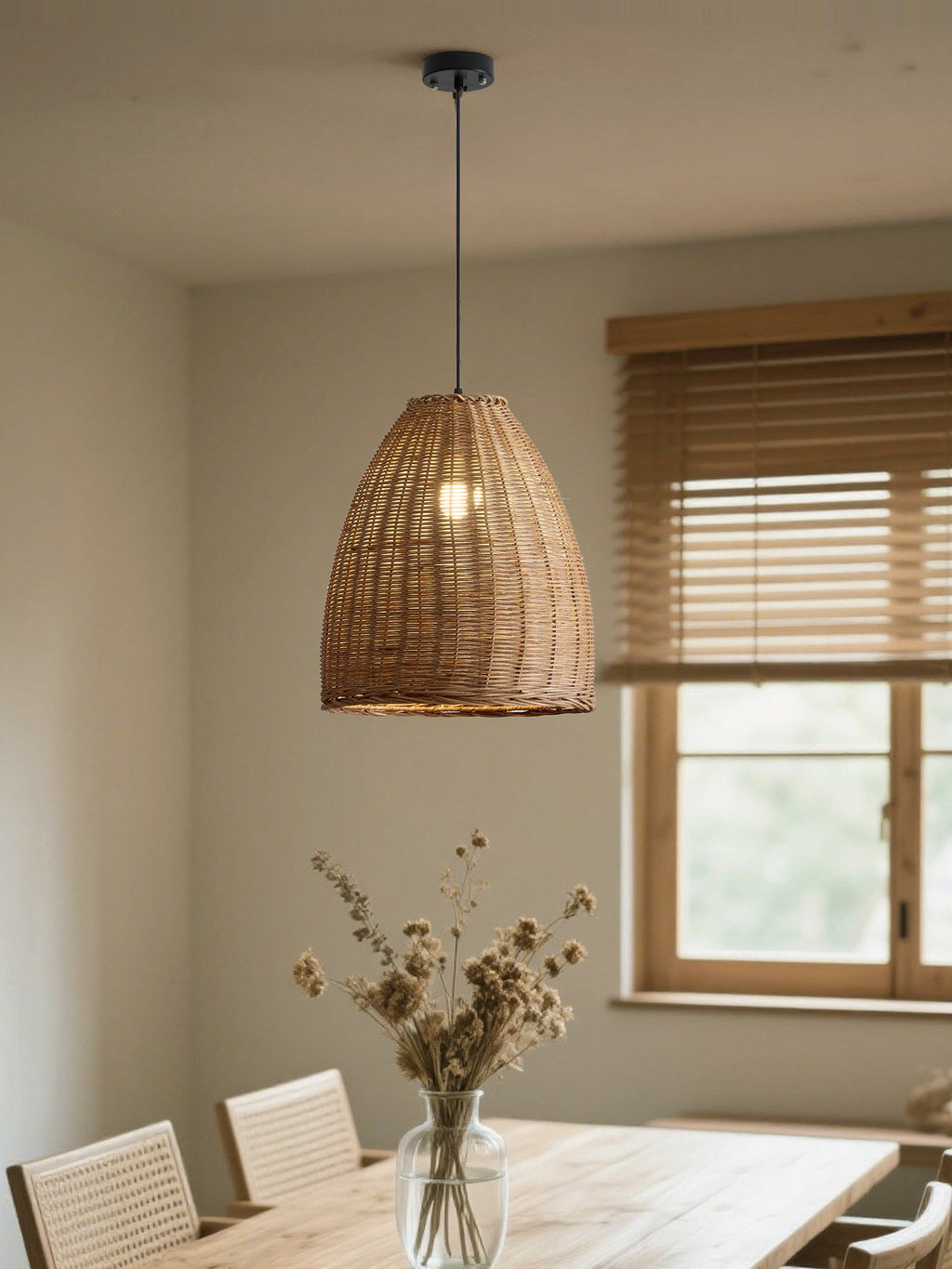 Dried flowers in a glass vase sit near a window with wood blinds, lit by this rattan Clérachic pendant.