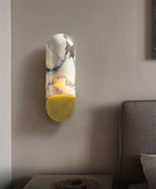 A grey sofa arm and round table with a small dish sit beneath the warm glow of the Clérachic marble cylinder wall light.