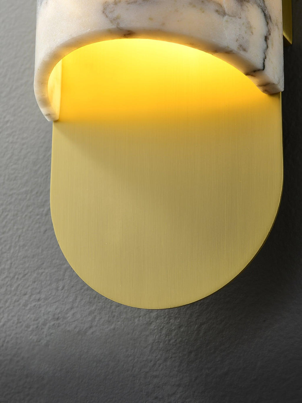 Clérachic marble cylinder wall light in extreme close-up, the curved brass base plate glowing warm beneath the marble.