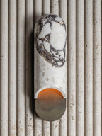 Clérachic marble cylinder wall light mounted on a fluted stone wall panel, its cylinder unlit and cool in tone.