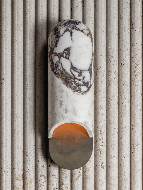 Clérachic marble cylinder wall light mounted on a fluted stone wall panel, its cylinder unlit and cool in tone.
