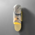 Clérachic marble cylinder wall light glowing warm on a plain grey wall, its brass base plate catching the light.