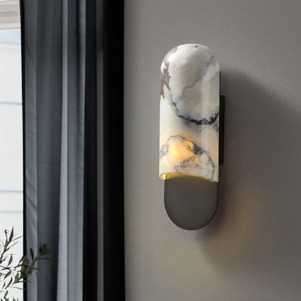 Clérachic marble cylinder wall light glowing warm beside a dark curtain and window, mounted on a plain grey wall.