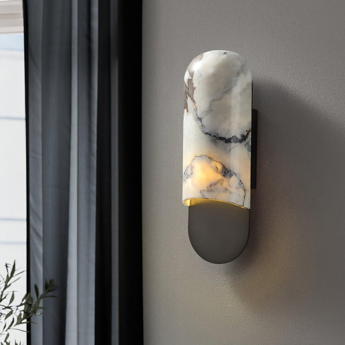 Clérachic marble cylinder wall light glowing warm beside a dark curtain and window, mounted on a plain grey wall.