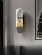 A dark mirrored panel reflects the Clérachic marble cylinder wall light's warm glow, a folded towel on the rail below.