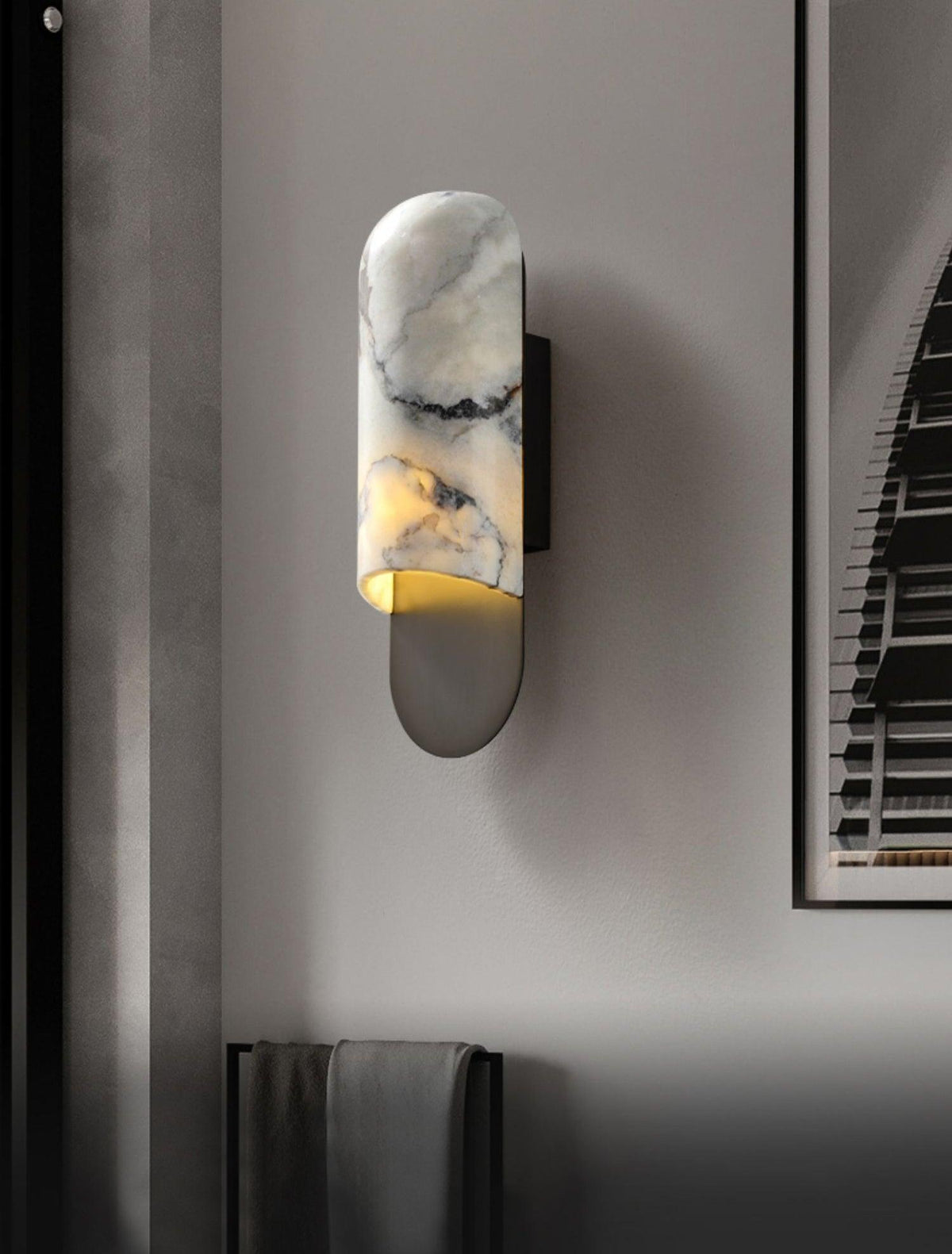 A dark mirrored panel reflects the Clérachic marble cylinder wall light's warm glow, a folded towel on the rail below.