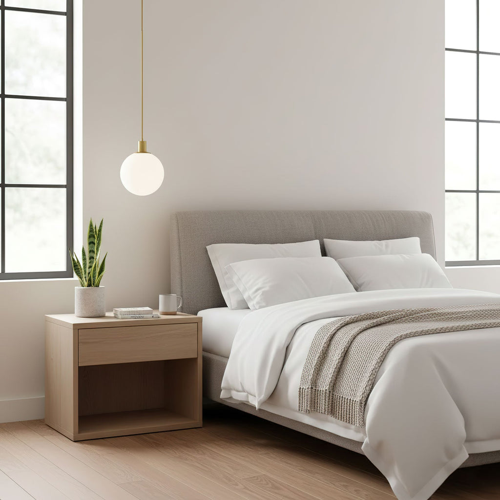 A single brass-and-white Clérachic sphere pendant hangs lit beside a bed with a grey headboard.