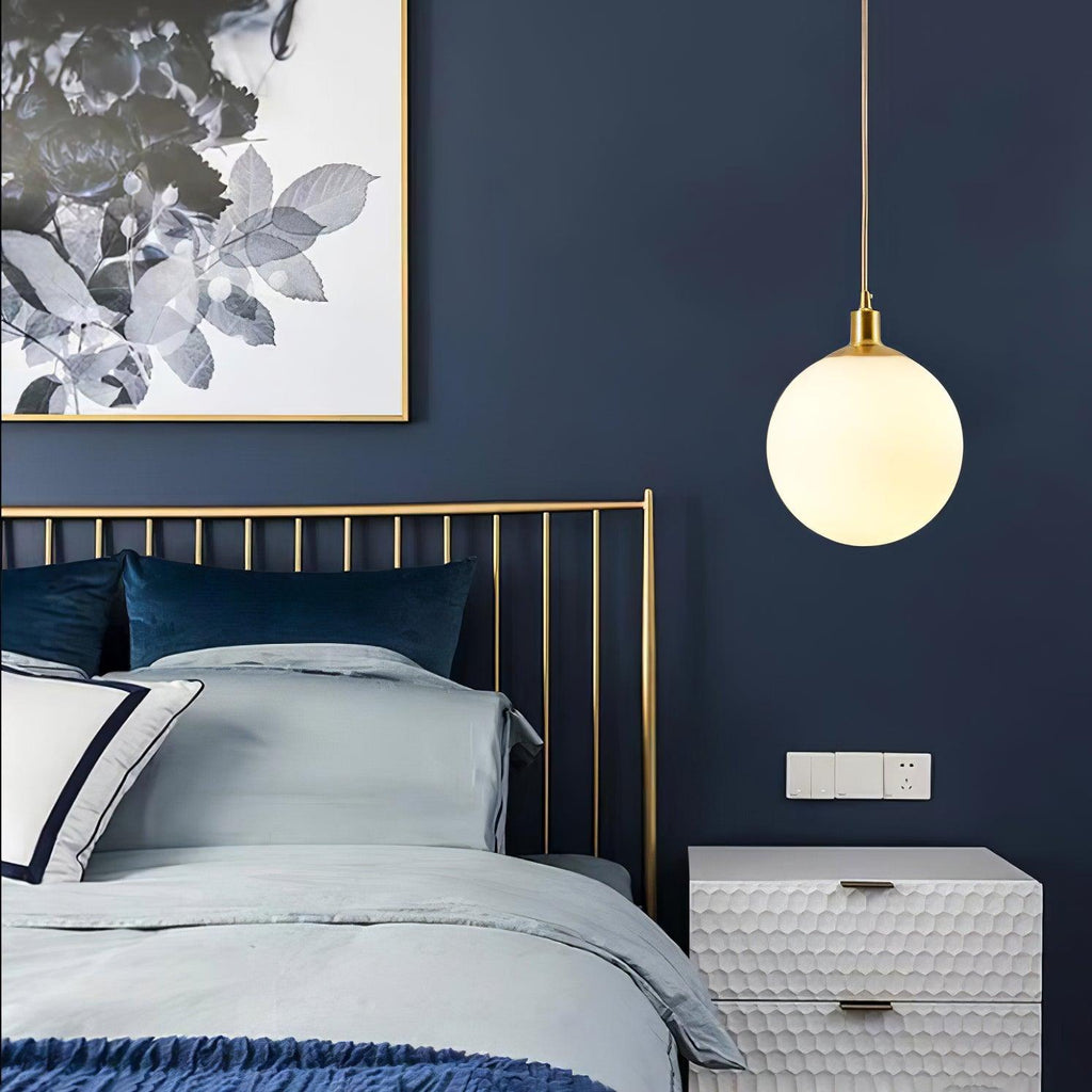 A single brass-and-white Clérachic sphere pendant hangs beside a brass bed frame on a navy wall.