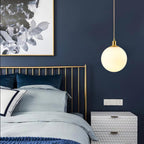 A single brass-and-white Clérachic sphere pendant hangs beside a brass bed frame on a navy wall.