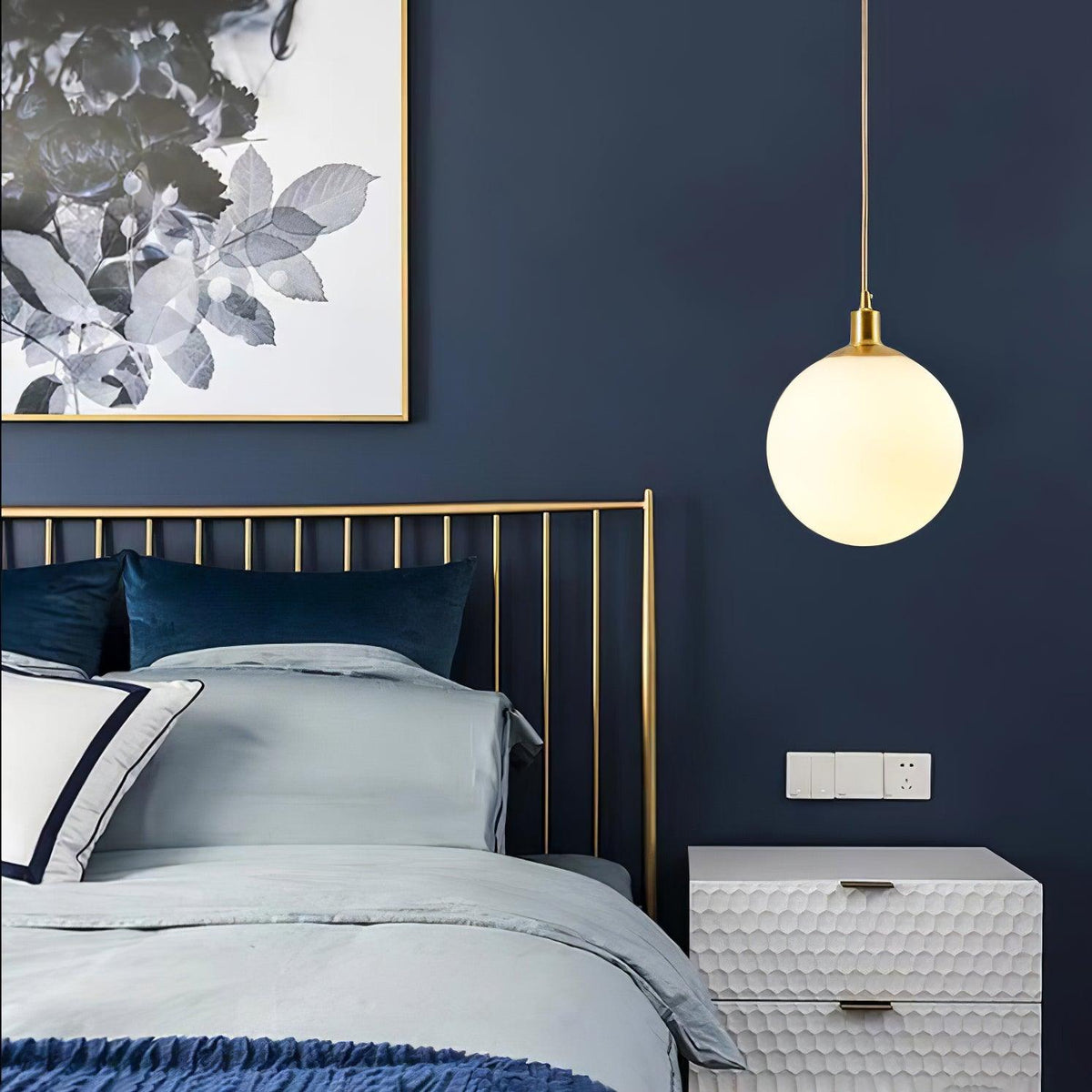 A single brass-and-white Clérachic sphere pendant hangs beside a brass bed frame on a navy wall.